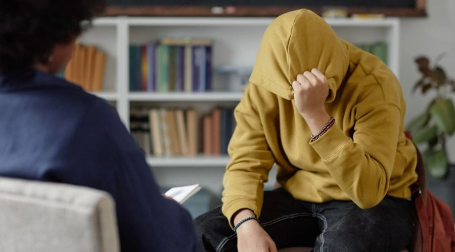 Image of a distressed teenage boy holding his hoodie over his face talking to a therapist