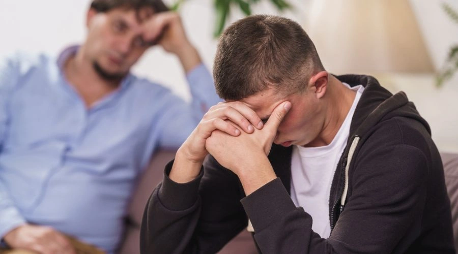Image of a frustrated, teenage male sitting in a sofa with his hands over his face as his father looks on