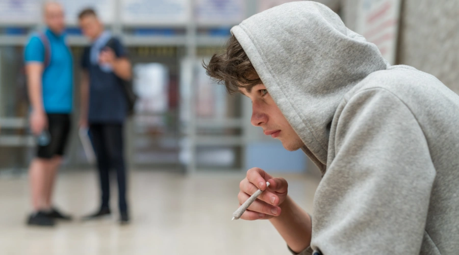 Image of a male teen wearing a hoodie and holding a weed cigarette with two males in the background - Long-term consequences of Adolescent substance abuse