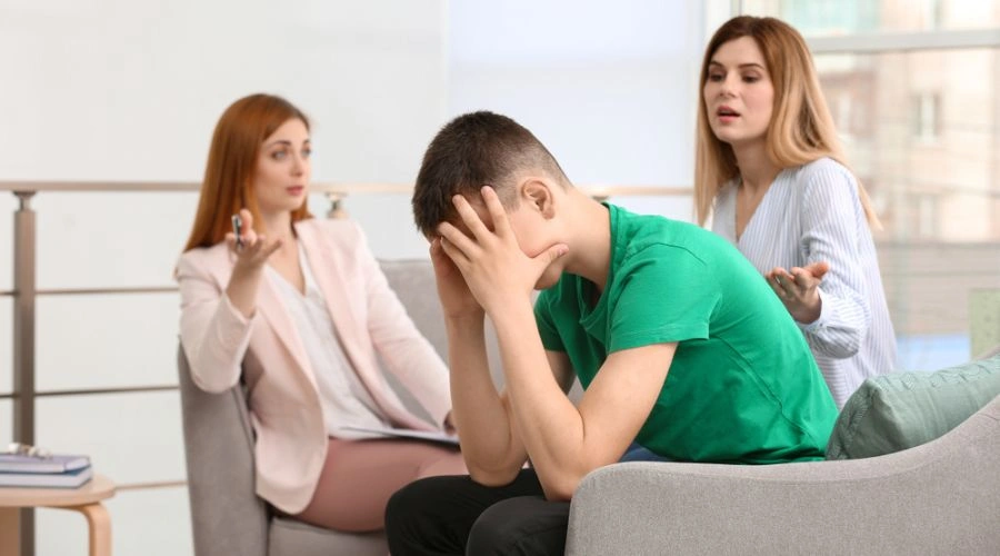 Image of a distraught teen male sitting in a chair with his hands on his face as his mother and therapist look on - Schizophrenia Treatment for Teens at Artemis