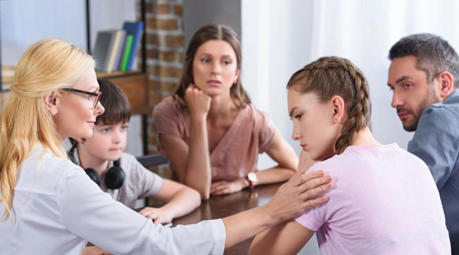 Image of a therapist holding her hand on a teenage females shoulder, as mother, father and younger brother look on - Structural Family Therapy for teens