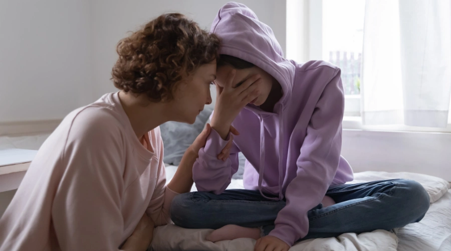Image of a worried mother comforting her depressed teen daughter, sitting on a bed with one hand over her face