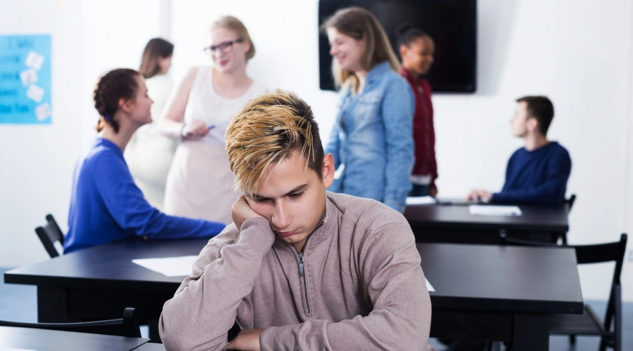 Image of Teen sitting alone in a classroom looking anxious while other students work together, symbolizing social struggles with schizotypal personality disorder