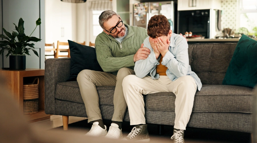 Image of a a father comforting his withdrawn teenager son, who is holding his hands over his face