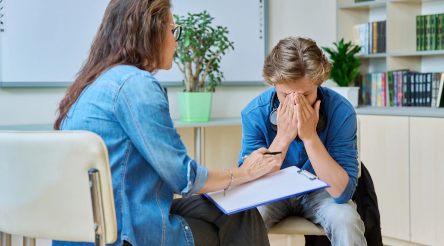Image of a male teen in therapy holding his hands over his face as a counselor looks on - 13 Mental Health Tips for Teens - Artemis Adolescent Healing Center