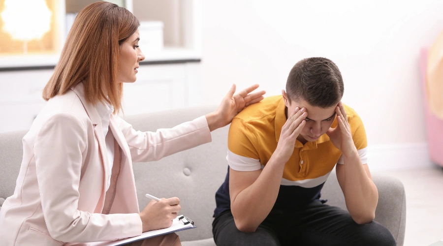 Image of a teen in therapy holding both of his hands on his forehead as a counselor looks on