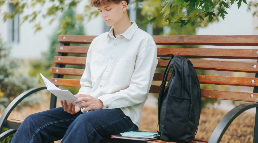 Image of a male teen sitting along on a park bench, reading a book