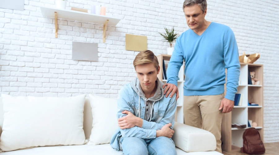 Image of father consoling his teenage son, who is sitting with his arms folded on a sofa - How to Help a Teenager with Mental Health Issues - Artemis Adolescent Healing Center