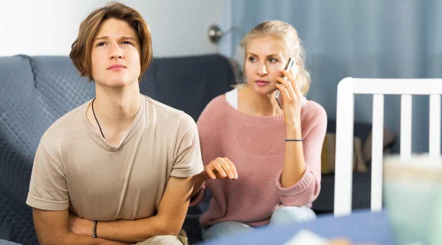 A concerned mother talking on the phone while sitting on a couch next to her distressed teenage son