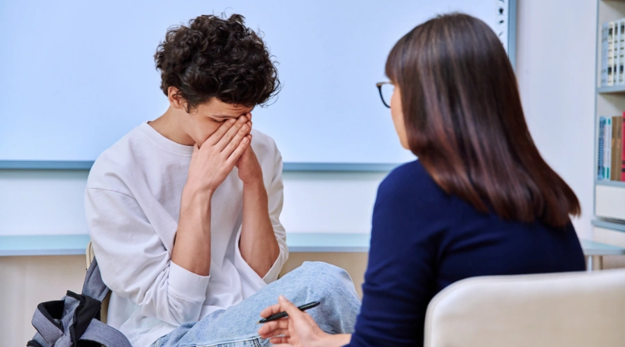 A distressed teenage boy wit covering his face with his hands while sitting across from a female therapist during a counseling session