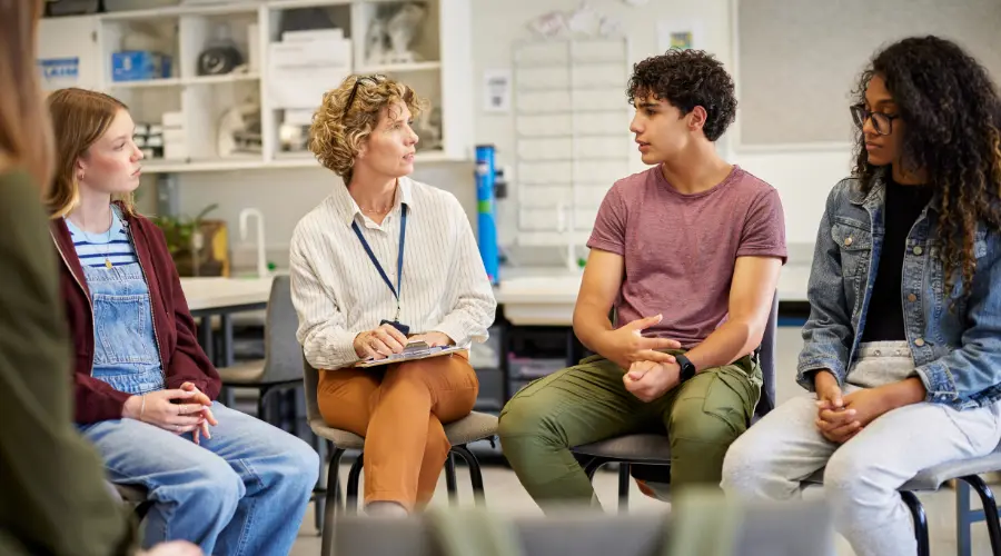 Image of a diverse group of teenagers seated in a circle with a female counselor taking notes, engaged in an open group therapy discussion
