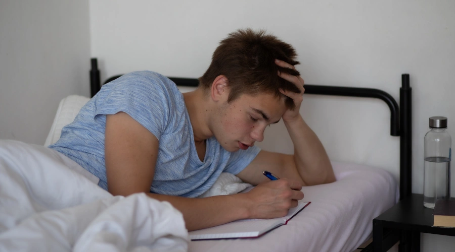 Image of a teenage boy lying on his bed writing in a journal with a pen